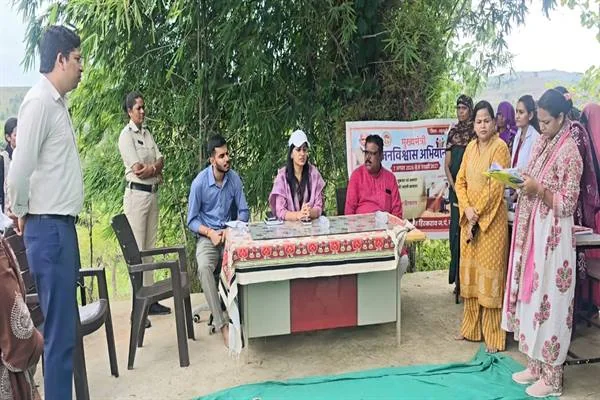 पेड़ों के नीचे लगे शिविर में मेज पर बैठे अधिकारी और सामने अपनी बात रखती महिलाएं