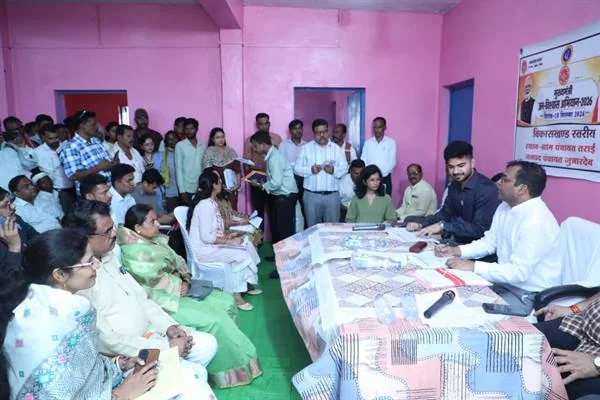 गुलाबी दीवारों वाले सभाकक्ष में मेज के दोनों ओर बैठे ग्रामीण और अधिकारी, पीछे खड़े लोगों का समूह तथा दीवार पर अभियान का पोस्टर
