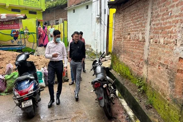 संकरी गली में खड़े दोपहिया वाहनों के बीच पैदल चलते अधिकारी और पीछे मौजूद स्थानीय लोग