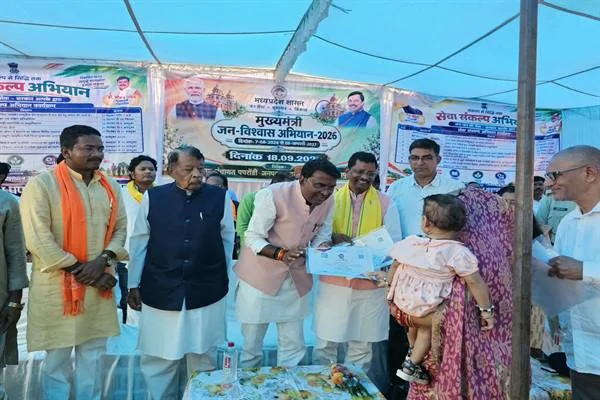 मंच पर मंत्री और अधिकारी एक महिला को दस्तावेज देते हुए, महिला की गोद में बच्चा है