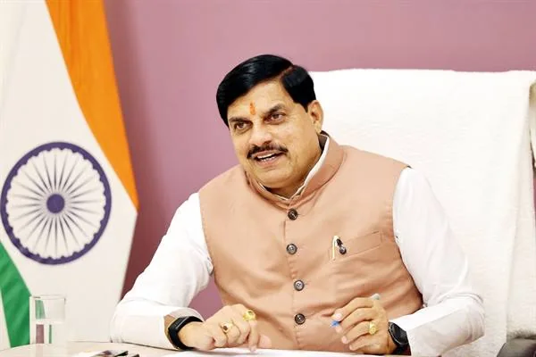 कुर्सी पर बैठे मुख्यमंत्री डॉ. मोहन यादव मुस्कुराते हुए बात कर रहे हैं, पीछे तिरंगा रखा है और सामने मेज पर पानी का गिलास है।